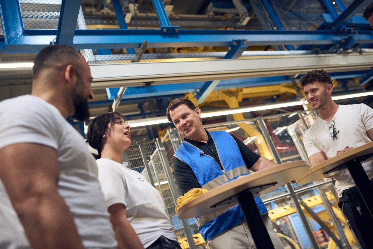 Jim Farley, wearing a blue safety vest over a darker blue polo shirt, speaks with Ford employees wearing white shirts.